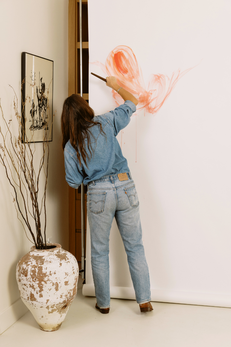 Artist in denim paints loose orange brushstrokes on a large sheet of white paper, using expressive movement and an oversized brush while exploring watercolor and masking fluid inspired techniques.
