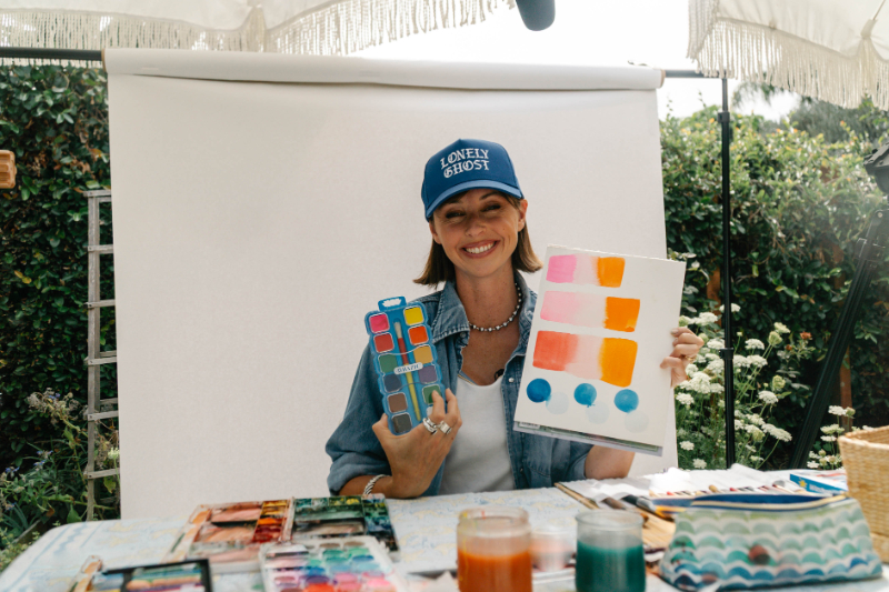Artist smiling at her painting table while holding a watercolor palette and a sheet of colorful swatches, demonstrating watercolor and masking fluid techniques with pink, orange, and blue washes.