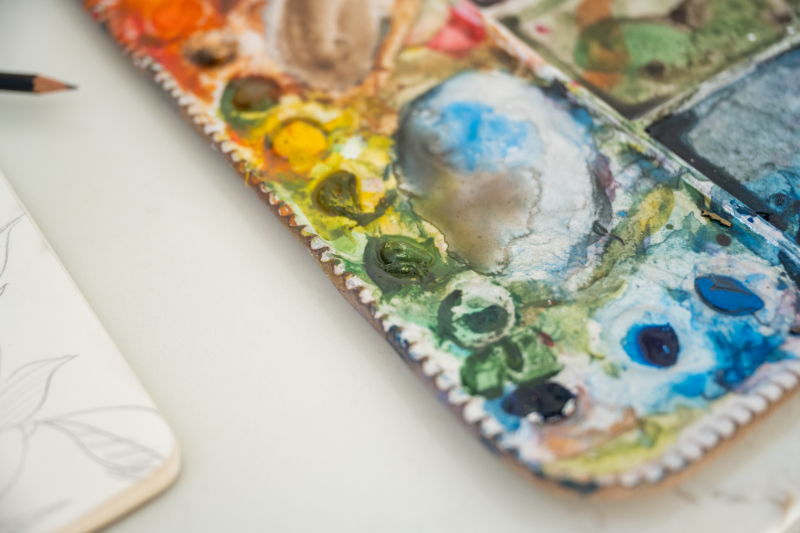 Close-up of a well-used watercolor palette filled with dried blue, green, yellow, and orange pigments beside a sketchbook, ready for experimenting with watercolor and masking fluid techniques.