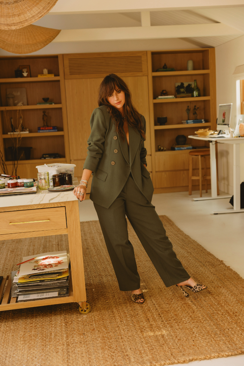 Artist wearing a dark tailored suit stands beside a worktable filled with paints and creative supplies in her bright home studio. The setting offers a personal look at the artist behind the work and her approach to art critique.