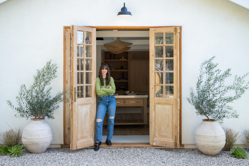 Artist stands with her arms crossed in the open doorway of a bright home art studio, framed by large potted trees and wooden doors. The welcoming creative space sets the scene for painting, learning, and thoughtful art critique.