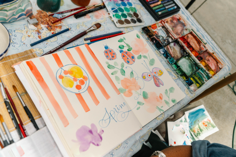 Open sketchbook filled with colorful flowers, leaves, insects, and orange stripes sits among brushes and watercolor palettes. The layered washes and transparent colors provide playful inspiration for practicing watercolor glazing.