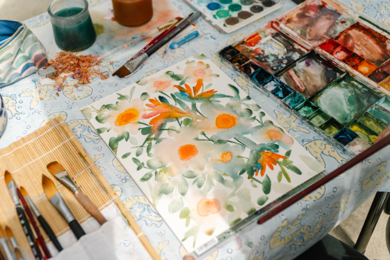 Loose watercolor painting of orange and peach flowers with soft green foliage sits beside brushes, palettes, and paint water. Transparent washes overlap throughout the floral study, showing the soft layered effects of watercolor glazing.