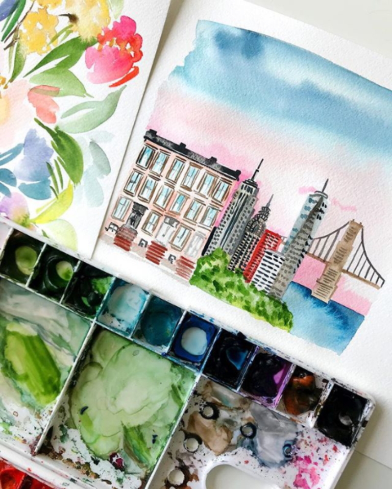 Watercolor cityscape features buildings, skyscrapers, a bridge, trees, and a softly blended blue and pink sky beside a floral painting and palette. The transparent layers of color show how watercolor glazing can build depth and atmosphere.