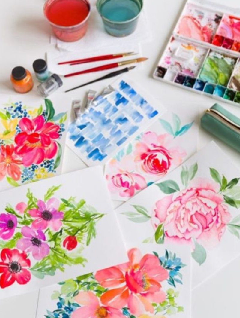 Collection of vibrant watercolor florals and blue studies arranged with brushes, paints, palettes, and colorful water cups. The overlapping washes and variations in color demonstrate creative possibilities for watercolor glazing.
