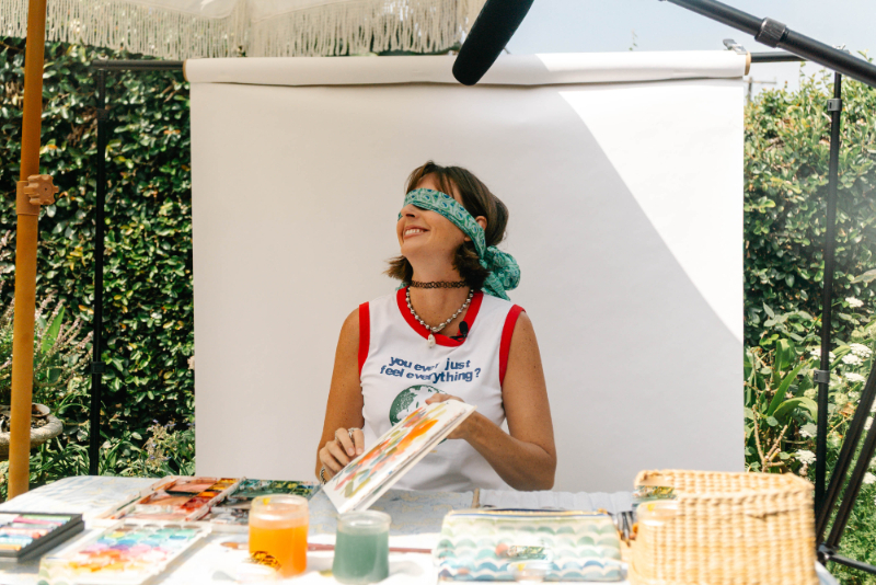Jenna Rainey smiles while blindfolded and holding a watercolor palette, exploring playful watercolor practice ideas that encourage creativity and letting go of perfection.